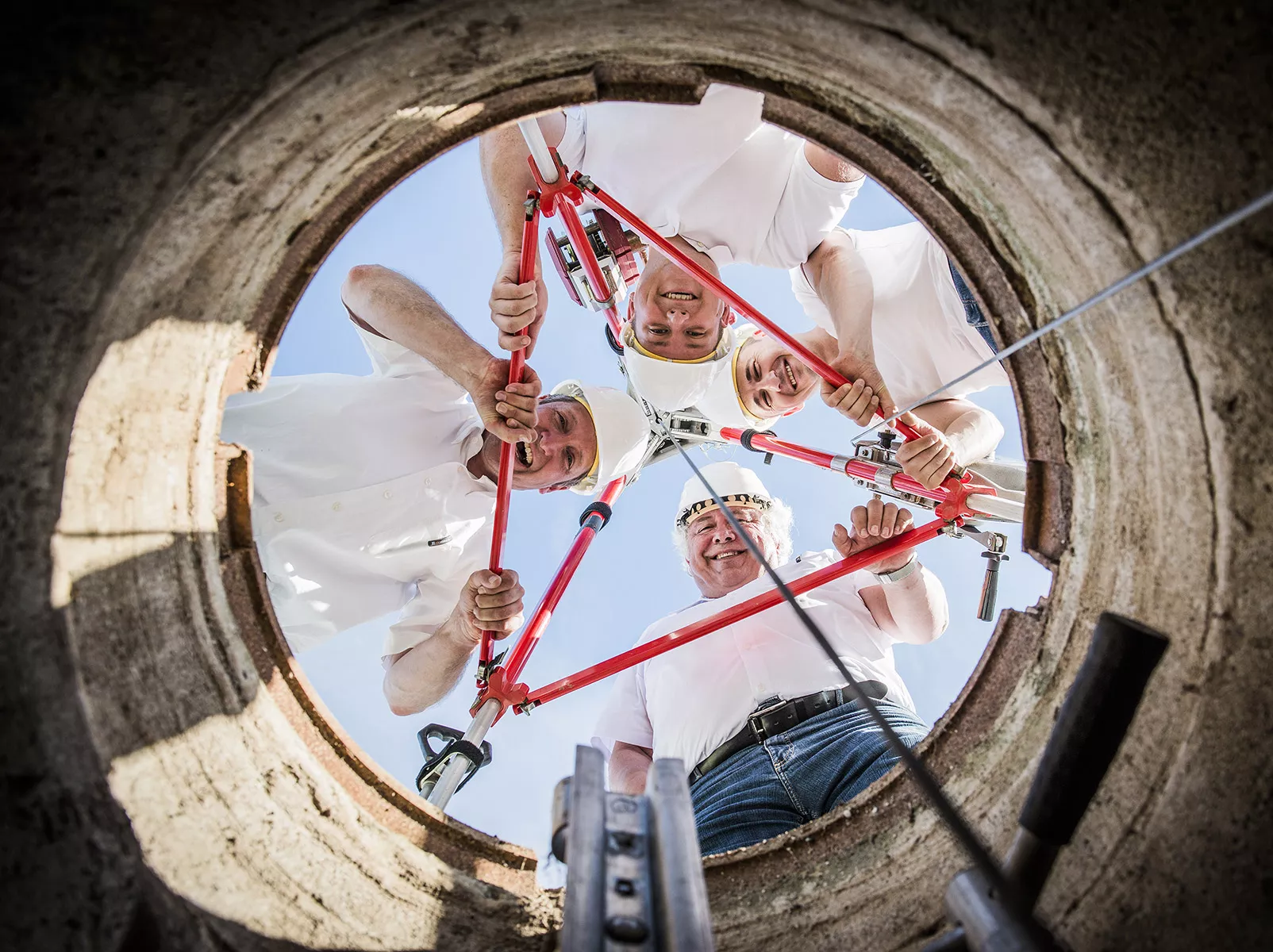 Blick, an einem Stahlseil hängend, nach oben durch einen Kanaldeckel auf 4 in die Kamera schauende Ingenieure